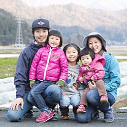 山脈と畑を背景に、肩を寄せ合いながらしゃがんで笑顔を向ける3人の子供と夫婦の写真