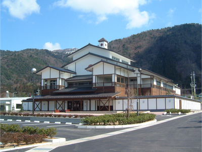 大きな山の前に白と木目が基調の大きな建物と駐車場が写っている写真(きららの湯)