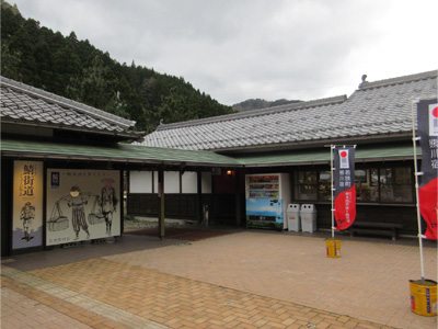 曇り空にL字に並んで建つ建物と、その前に自動販売機や黒と赤ののぼり旗が立っている様子が写った写真(道の駅 熊川宿)
