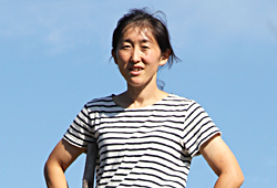 雲ひとつない青空を背景に腰に手を当てている八代恵里さんの上半身アップの写真