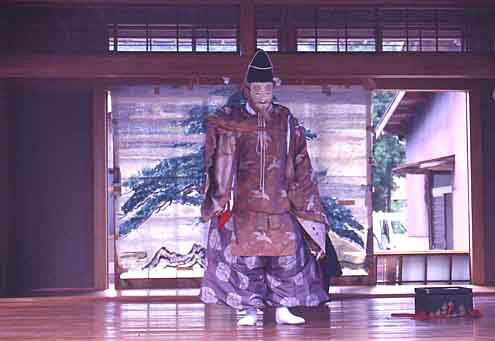 木の絵が描かれた広めの掛け軸がかけられた板の間で茶色っぽい着物と紫っぽい袴を履き能面をつけた人がいる写真