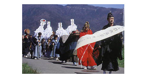 神事の様子で、黒や赤の浄衣などを着た人々が手に旗やおおぬさを持ち、列を成して歩いている様子の写真