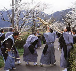 肩衣がついた袴を着た数人の男性がロープで括った木材のロープを上に引っ張って木材を上に浮かせている写真