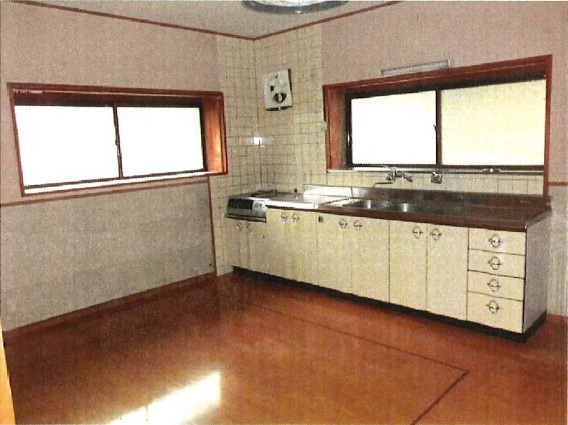 壁付のキッチンと換気扇、ダイニング部分はフローリング、大きな腰窓が2つ写っている民家の室内の写真