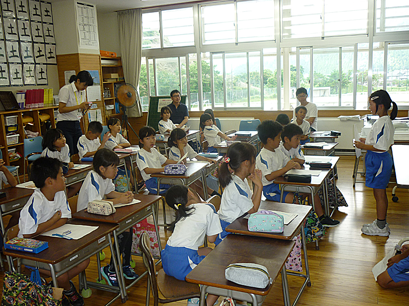 教室で1人の女の子が前で発表しているのを、他の生徒や先生が見ている写真