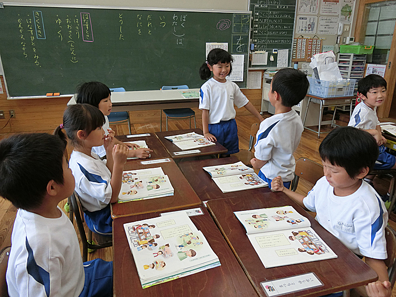 教室で6人の生徒が向かい合ってグループになって座り、机の上に教科書を開いて話し合っている写真