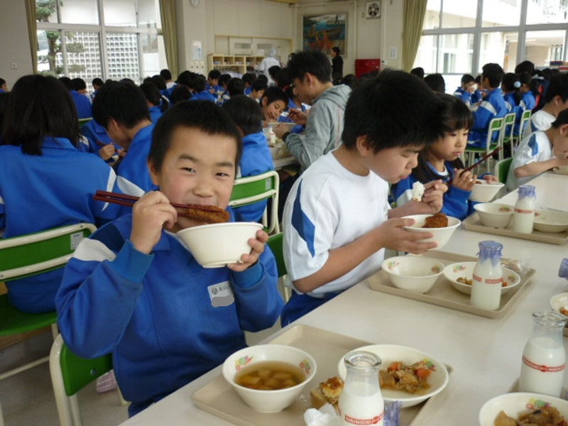 たくさんの生徒が給食を食べていて、1人の男の子が揚げ物を頬張りながらこちらを見てにっこりしている写真