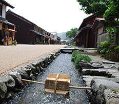 街道と家屋の間に石積みの用水路があり、木製の小さな水車が用水路に渡されている写真