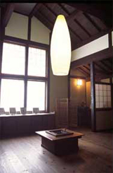 天井が高く大きな窓から光が差し込み、木製のフローリング、真ん中に囲炉裏、その上には天井から吊り下がっている大きなデザイン照明のある部屋の写真