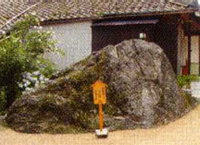 古い家屋の前にとても大きく、どっしりとした岩がありその前には木製の看板が建てられている写真