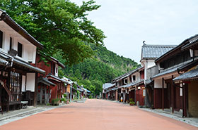 オレンジ色の道を中央に左右に家屋が並び、奥に青青とした山が写っている写真