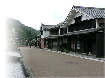 中央に赤褐色の大きな街道があり、左側には用水路、右側には古い家屋が立ち並ぶ写真